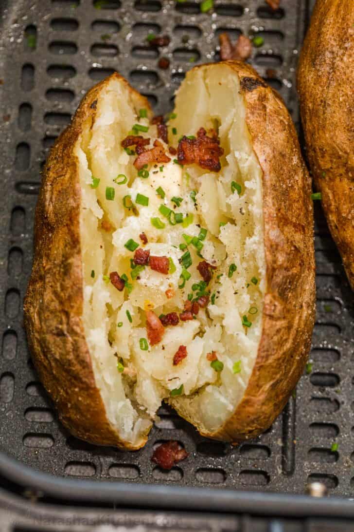 Easy and fail-proof recipe for Air Fryer Baked Potatoes. Potatoes in the air fryer have crisp salty skin and a soft and fluffy center. Baked potato loaded with butter, bacon, and green onion in an air fryer basket.