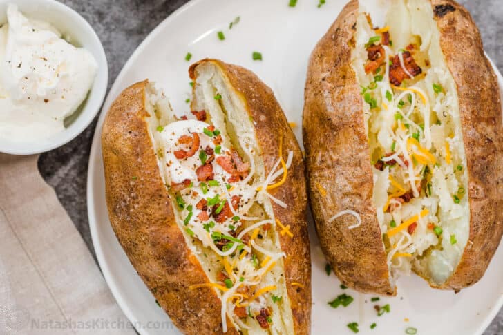 A classic baked potato recipe that is made in an air fryer that gives you a crispy skin and a soft and fluffy potato. Two fully loaded with baked potatoes on a white plate next to sour cream.