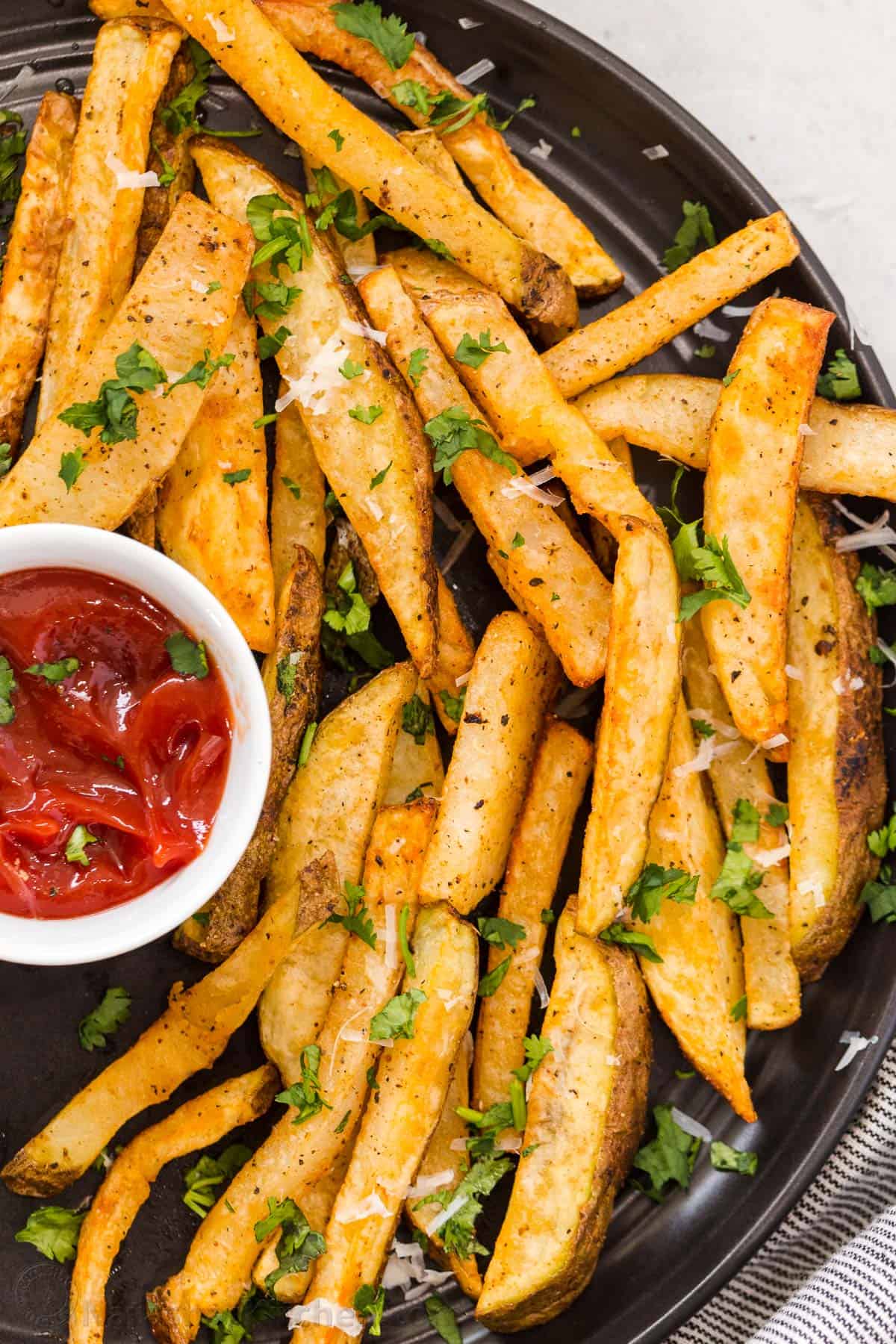 A SIMPLE recipe for air fryer French fries that are crispy, crunchy and seasoned to perfection. Homemade air fryer French fries on a black plate with a bowl or ketchup.