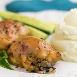 A plate of baked mushroom stuffed chicken thighs besides mashed potatoes and cucumbers