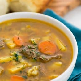 A white bowl of beef, barely and pickle soup with bread in the background