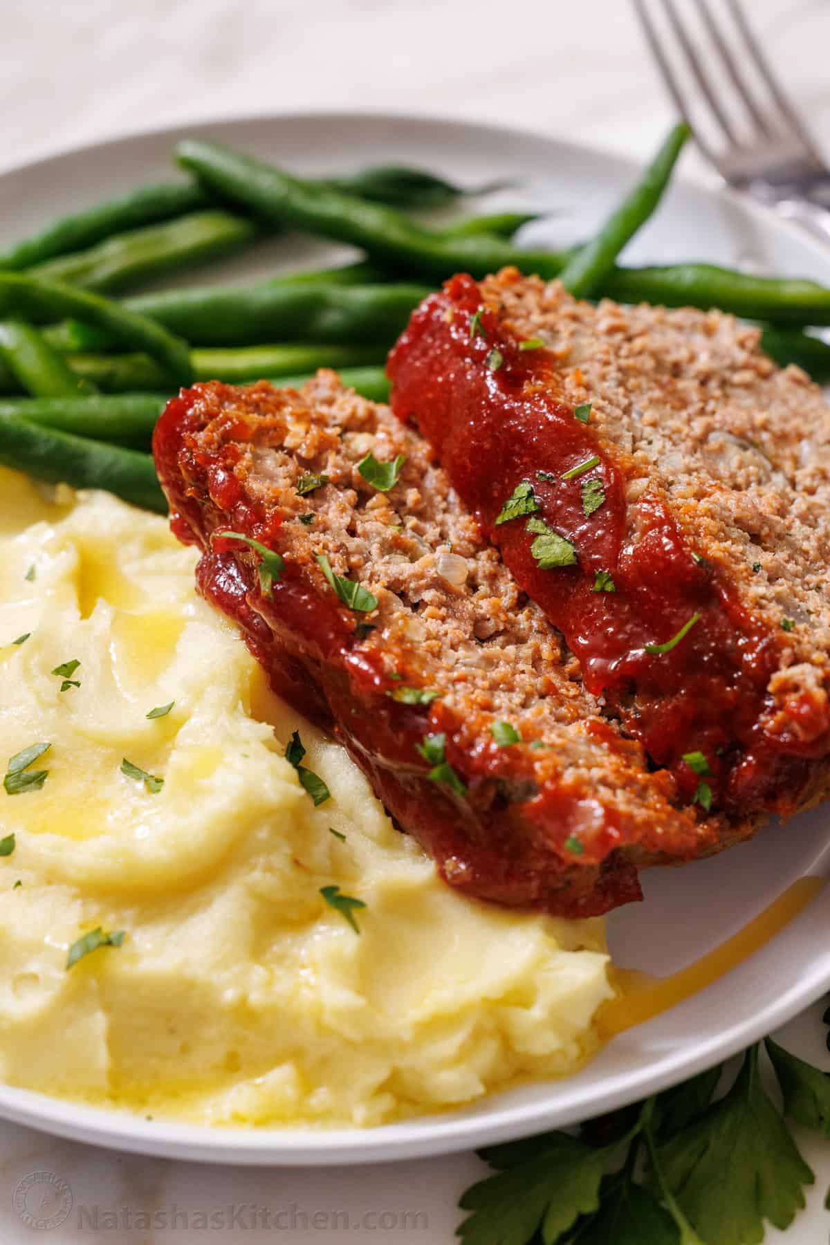 Amazing homemade Meatloaf Recipe. The meatloaf is so tender and juicy on the inside with a sweet and tangy sauce that glazes the meatloaf and adds so much flavor! #meatloaf #meatloafrecipe #howtomakemeatloaf #glazedmeatloaf #meatloafsauce #bestmeatloaf #natashaskitchen #groundbeef Meatloaf Served with mashed potatoes and green beans on a plate