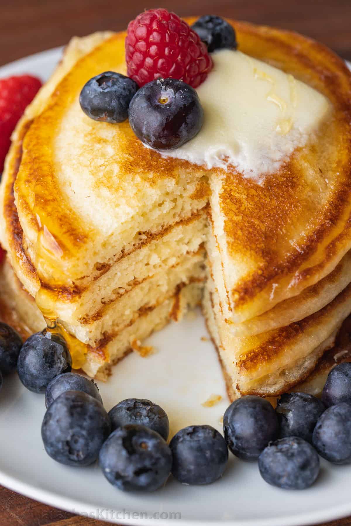 Fluffy Homemade Buttermilk Pancakes are the perfect breakfast. This pancake recipe is quick and easy to make (done in less than 30 minutes!) Stack of buttermilk pancakes with slice cut out topped with butter, syrup blueberries and a raspberry