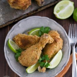 A plate of breaded baked chicken drumsticks garnished with lime wedges and cilantro