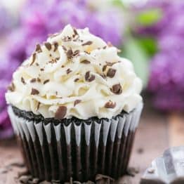 A close up of a dark chocolate cupcake with white chocolate frosting with flowers behind it