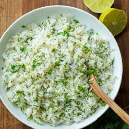 Cilantro lime rice in a large white serving bowl with a wooden spoon and slice limes