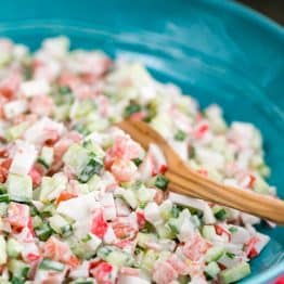 A blue bowl of crab salad with cucumber and tomato with a wooden spoon in it