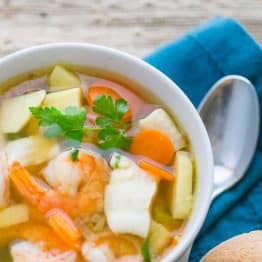 A white bowl of fish and shrimp soup, ukha, with a spoon, and bread bun besides it