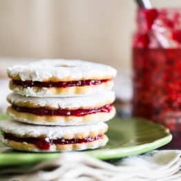 A close up of three jam-filled sandwich cookies on a plate with a jar of jam in the background