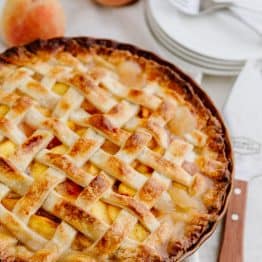 A peach pie with a spatula, plates, forks and fresh peaches beside it