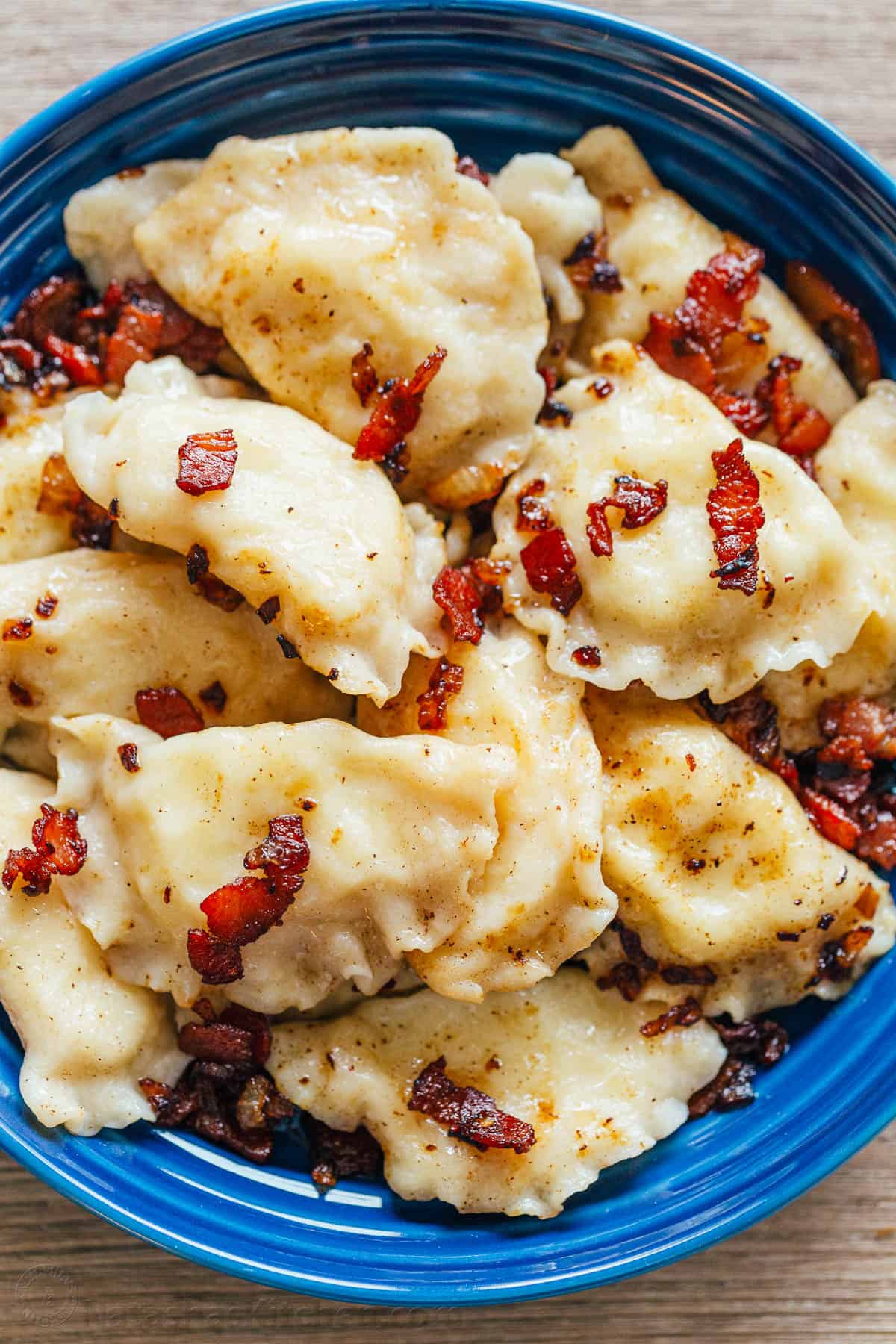 Pierogi filled with cheesy potatoes and served with crisp bacon, melted butter and sour cream. Homemade Pierogi is the ultimate comfort food! Cooked pierogies on a blue plate with bacon