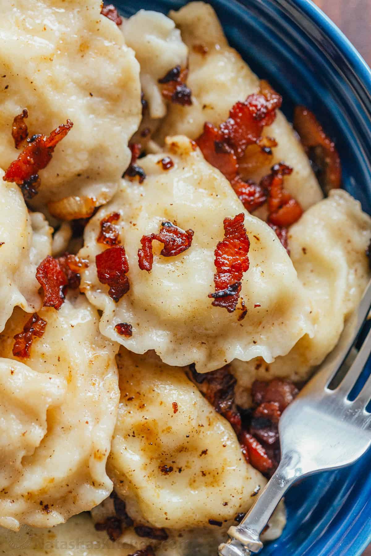 Pierogi filled with cheesy potatoes and served with crisp bacon, melted butter and sour cream. Homemade Pierogi is the ultimate comfort food! The best pierogi recipe on a blue plate with bacon