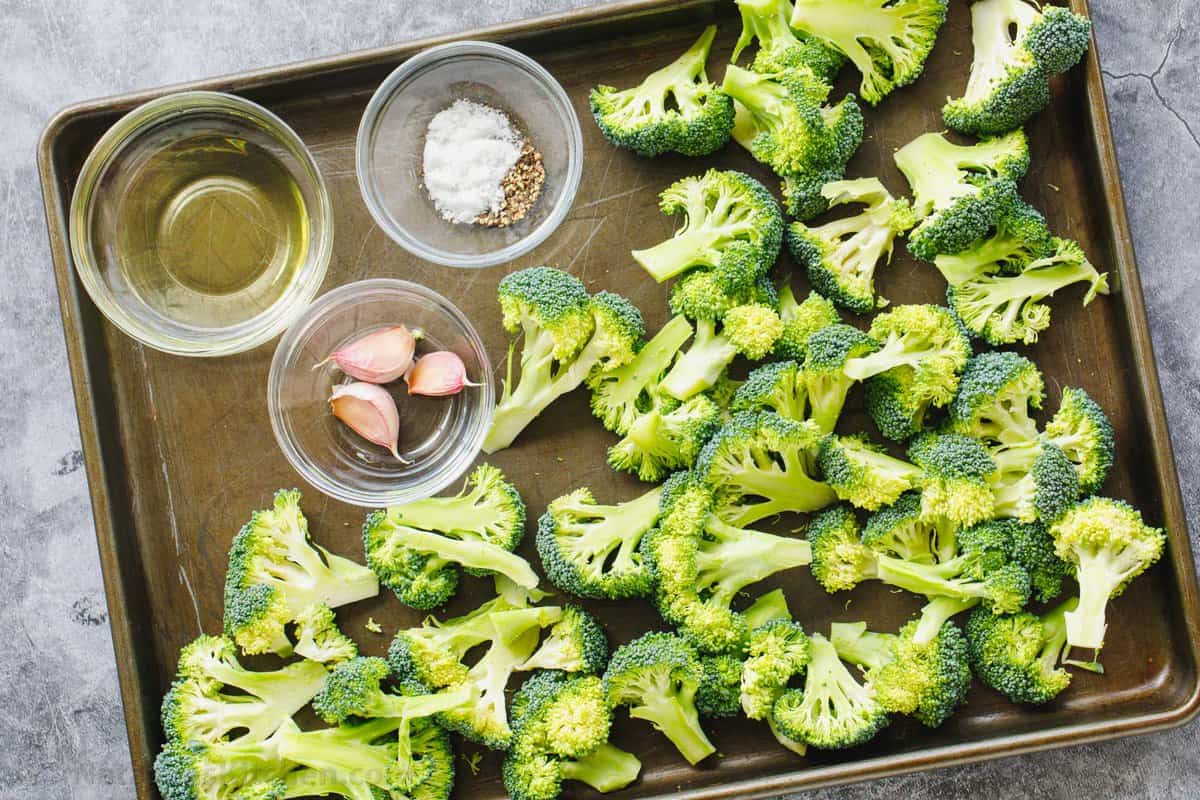 Raw broccoli florets with oil, garlic and seasonings in ramekins on a baking sheet.