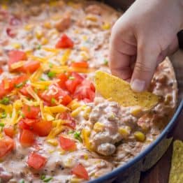 Sausage Queso Dip in pan topped with tomato, green onion, and cheese with a hand dipping a chip in it.