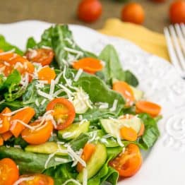 A plate of spinach salad with tomatoes and cucumbers in the background and a fork beside the plate