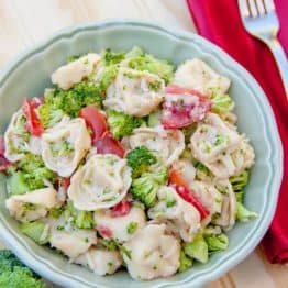 A bowl of tortellini salad with red, cloth napkin with a fork on top beside it