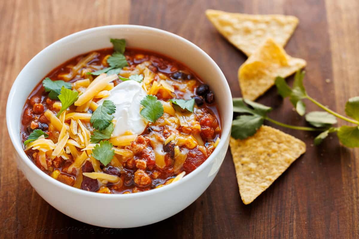 white bowl of turkey chili with cheese and sour cream on top and corn chips on the side